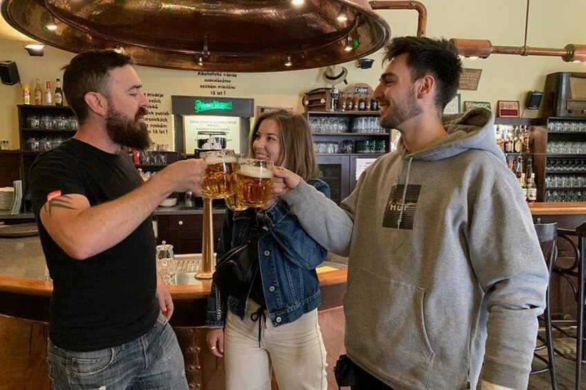 Tres personas brindando con vasos de cerveza en un bar con cañerías de cobre y estantes llenos de botellas. 3