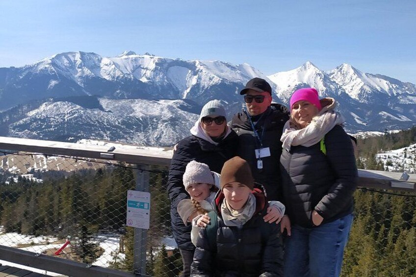 Eine Gruppe von Personen steht auf einer Aussichtsplattform, im Hintergrund schneebedeckte Berge. 1