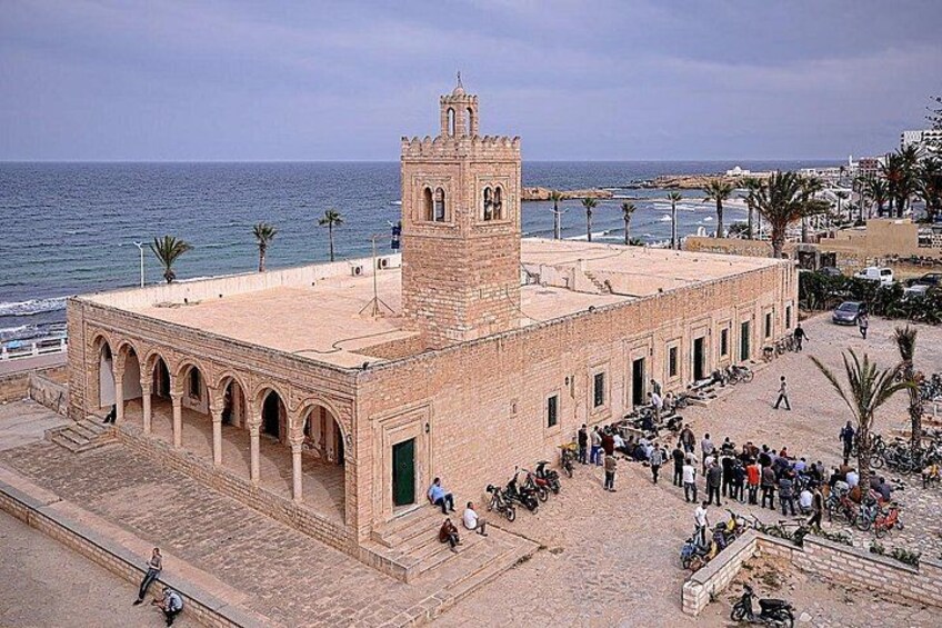 Un bâtiment historique doté d’une tour, une zone pavée où l’on peut voir des piétons et des vélos, et une vue sur la côte. 5