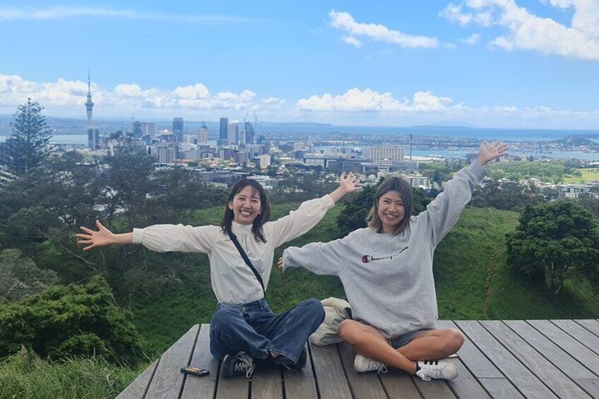 Two people sitting on a wooden deck with arms outstretched, overlooking a cityscape with a prominent tower. 4