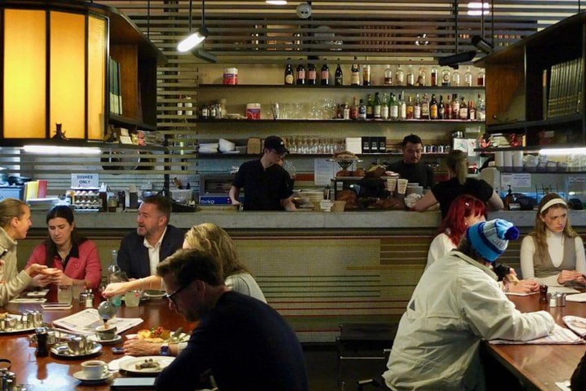 A restaurant interior with patrons dining, a bar area with bottles, and a counter where a server is attending to customers. 4