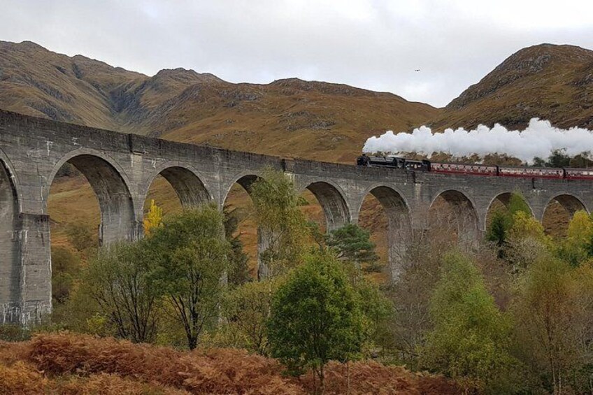 A train on a stone viaduct with arched supports, surrounded by trees and hills. 1