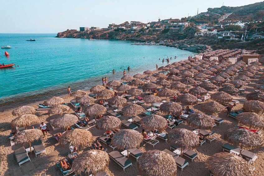Een strand met rieten parasols en ligstoelen, een heldere blauwe zee en gebouwen op de achtergrond. 3