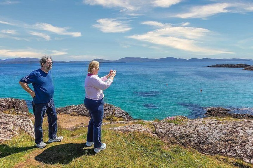 Deux personnes se tiennent sur une côte rocheuse, avec vue sur la mer et les montagnes au loin. 2