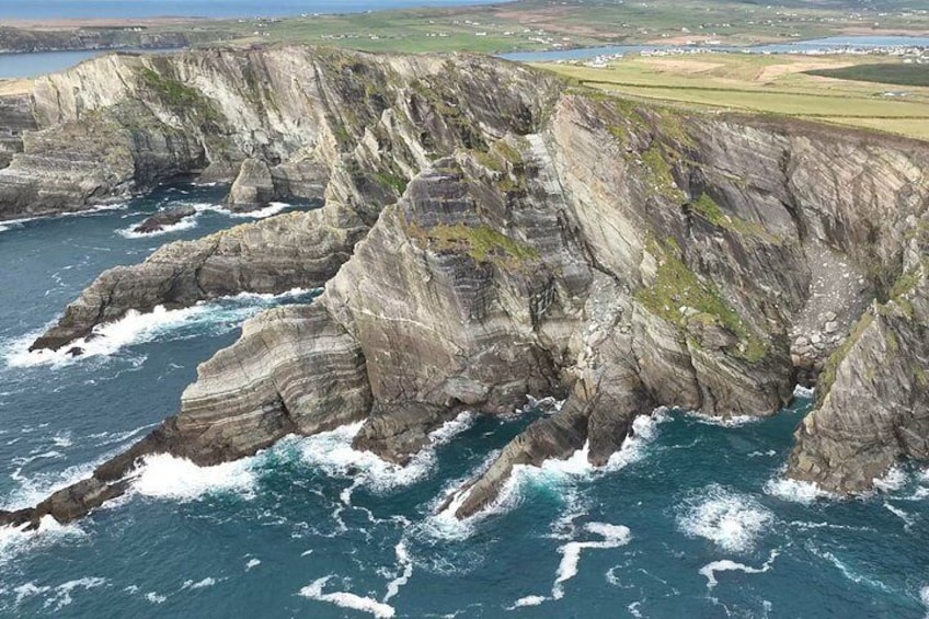 Une falaise côtière aux couches rocheuses stratifiées, où les vagues s’écrasent contre la base, et d’où l’on aperçoit au loin un paysage côtier. 5