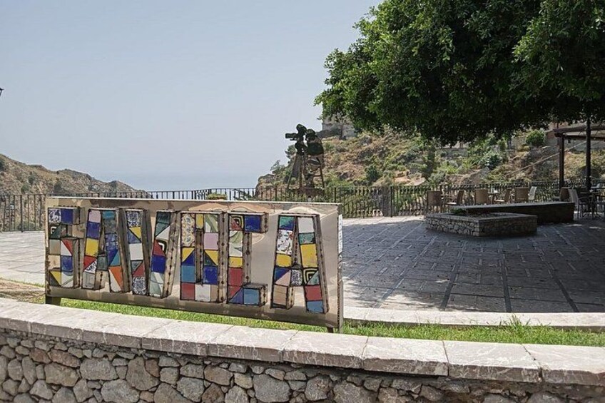 A mosaic wall with geometric patterns, a stone wall, and a paved area with a statue and seating. 5