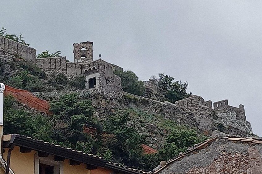 A historic stone fortress with battlements and a watchtower, surrounded by vegetation and modern buildings. 2