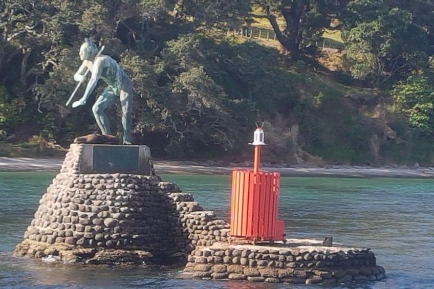 A statue on a stone pedestal by the water, a red cylindrical structure, and a rocky shoreline with trees in the background. 2