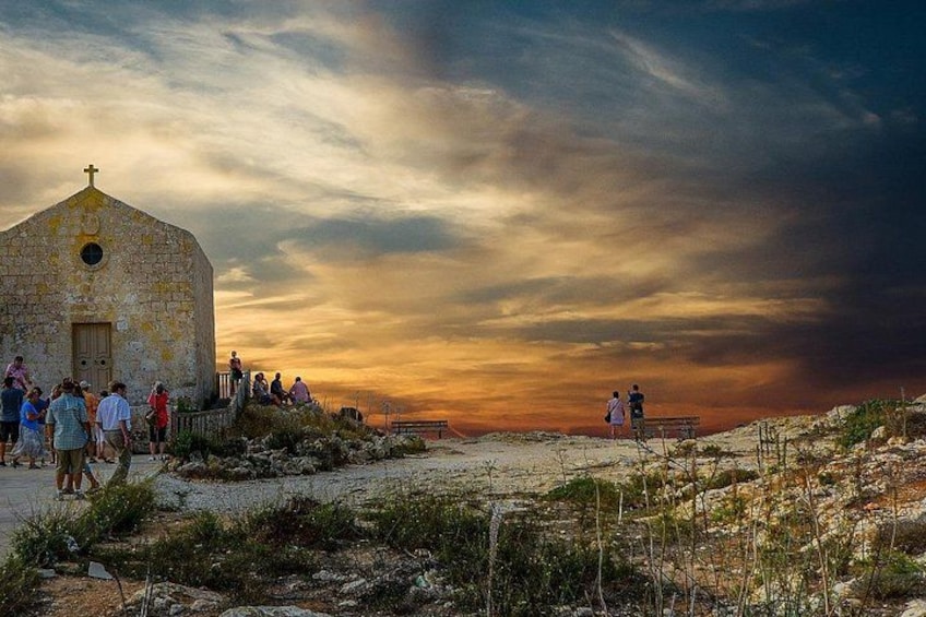 A stone church with a cross on top, people gathered around, and a sunset in the background. 2