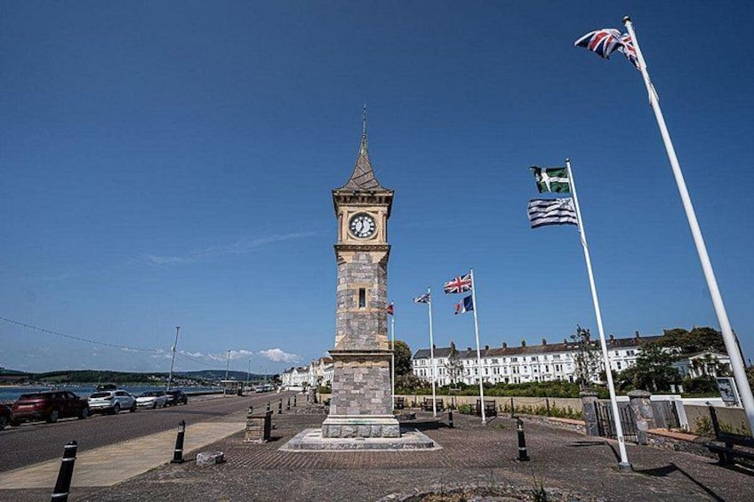 A clock tower with flags, a paved area, and a building in the background. 5