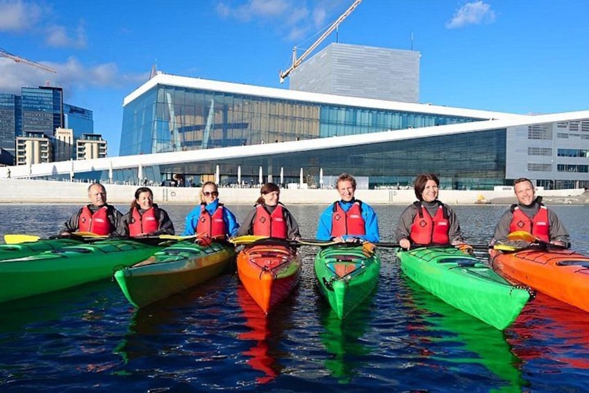 Seis personas en kayaks alineadas sobre un cuerpo de agua, con un edificio moderno al fondo. 3