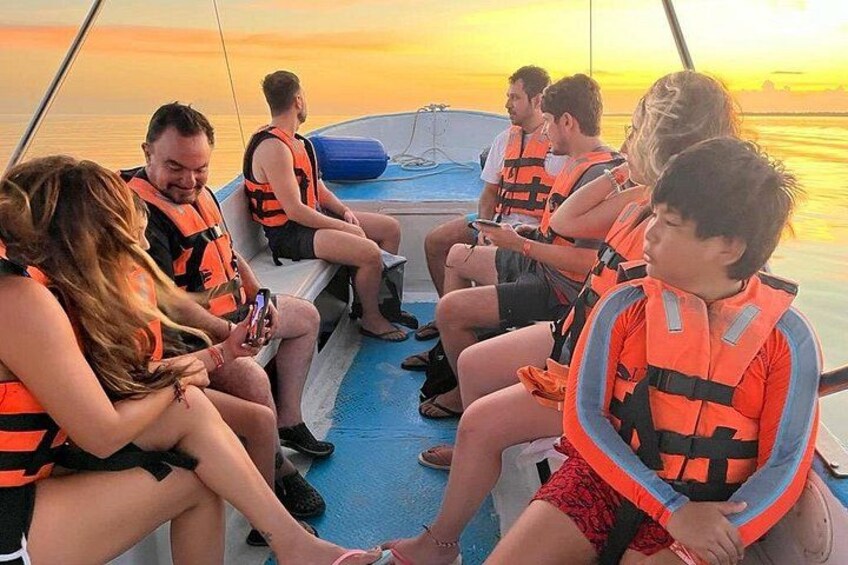 Een groep mensen in reddingsvesten op een boot, met een zonsondergang op de achtergrond. 1