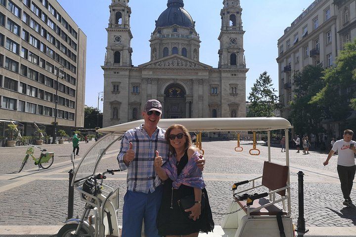 Un couple pose devant un bâtiment historique, avec un vélo et une charrette au premier plan. 2