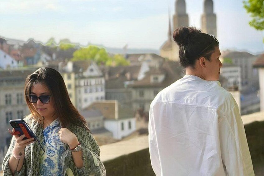 Two people, one using a smartphone, overlooking a cityscape with historic buildings and a prominent tower. 1