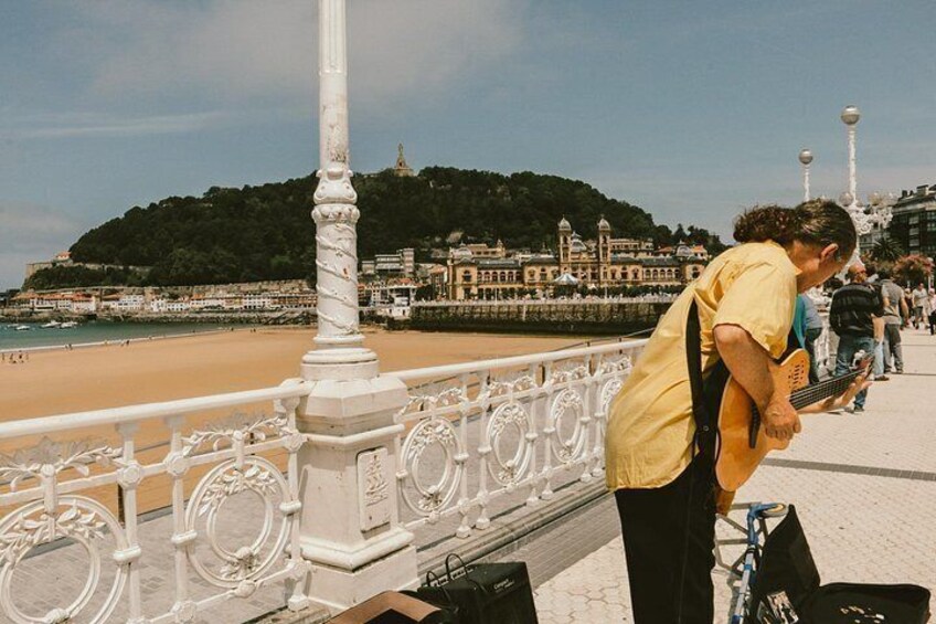 A person playing a guitar on a white ornate railing by a beach with a hill in the background. 2