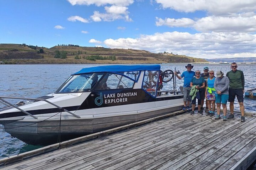 Een boot genaamd 'Lake Dunstan Explorer' ligt aan een houten steiger, waar een groep mensen staat. 3