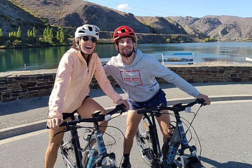 Twee personen op fietsen langs een meer, met bergen op de achtergrond. 5