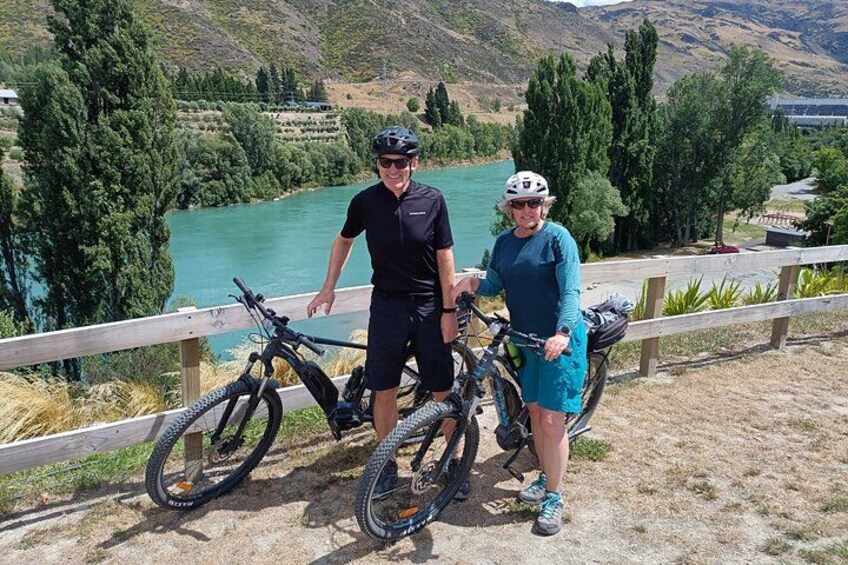 Twee personen staan naast fietsen op een houten terras met uitzicht op een meer, bomen en heuvels op de achtergrond. 4