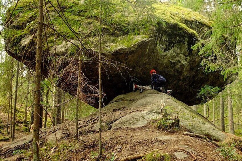 A person sitting on a large rock in a forested area with trees and a moss-covered rock formation. 2