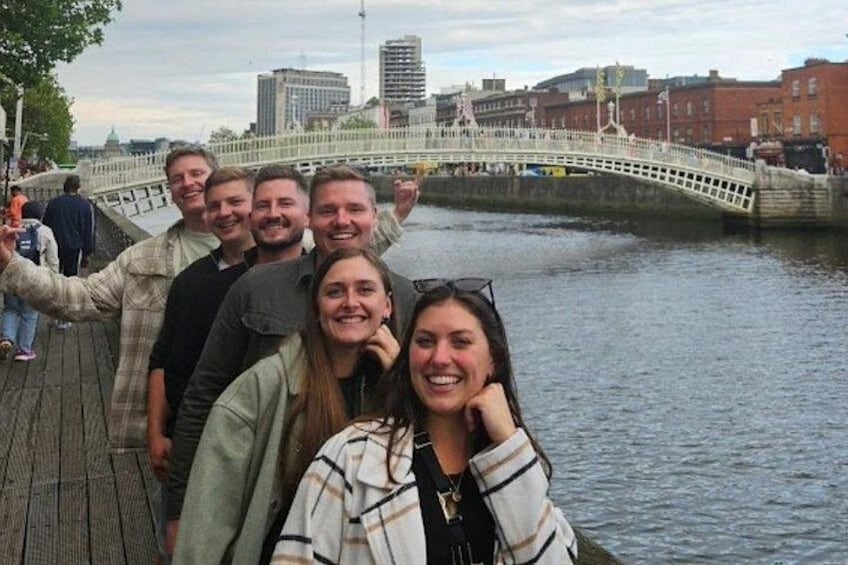 Eine Gruppe von Menschen auf einem hölzernen Steg entlang eines Flusses, im Hintergrund eine Brücke. 1