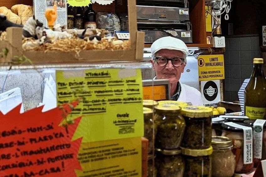 Una persona dietro un bancone con diversi prodotti alimentari e vasetti. Si notano cartelli con del testo e un cartello giallo di avvertimento. 4