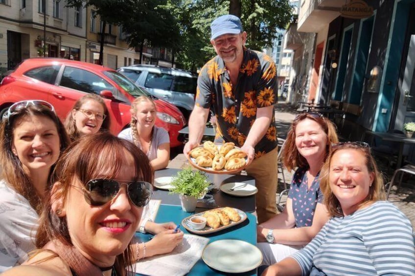 A group of people sitting at an outdoor table with food, a street with parked cars, and buildings in the background. 1