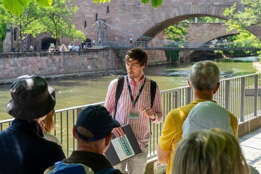 Une personne portant une chemise à carreaux et un sac à dos, debout près d’une rivière, avec un pont en arrière-plan. 3