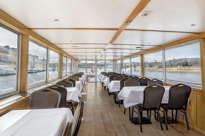 A dining area with tables and chairs on a boat, large windows along the sides, and a view of a river and buildings. 2
