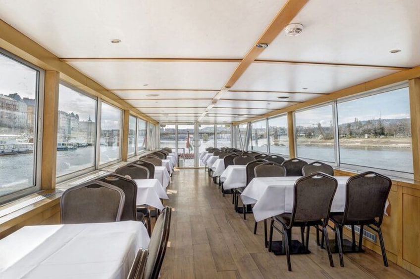 Comedor en un barco con mesas y sillas, ventanas amplias y vista al agua y la ciudad. 2