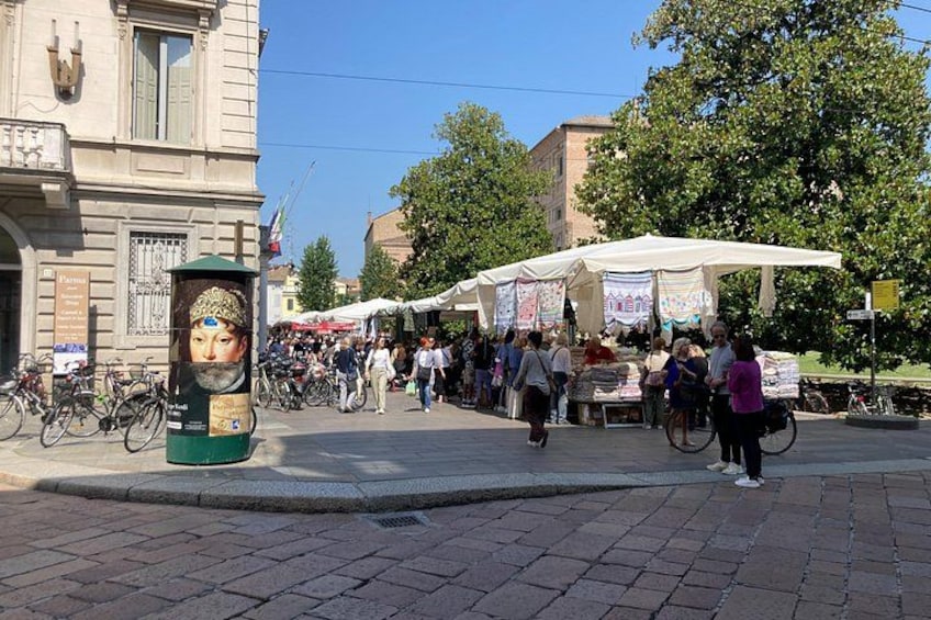 Un marché animé en plein air, où l’on peut voir des gens faire leurs emplettes et des vendeurs installés sous des tentes, un bâtiment historique et des vélos garés à proximité. 1