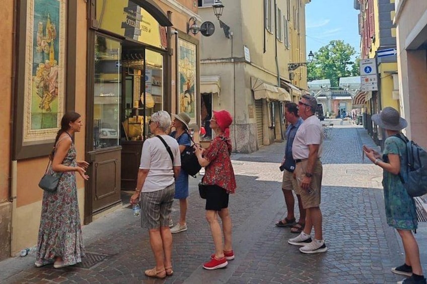 Un gruppo di persone in piedi su una strada acciottolata, un negozio con vetrine in vetro e un edificio con un cartello. 1
