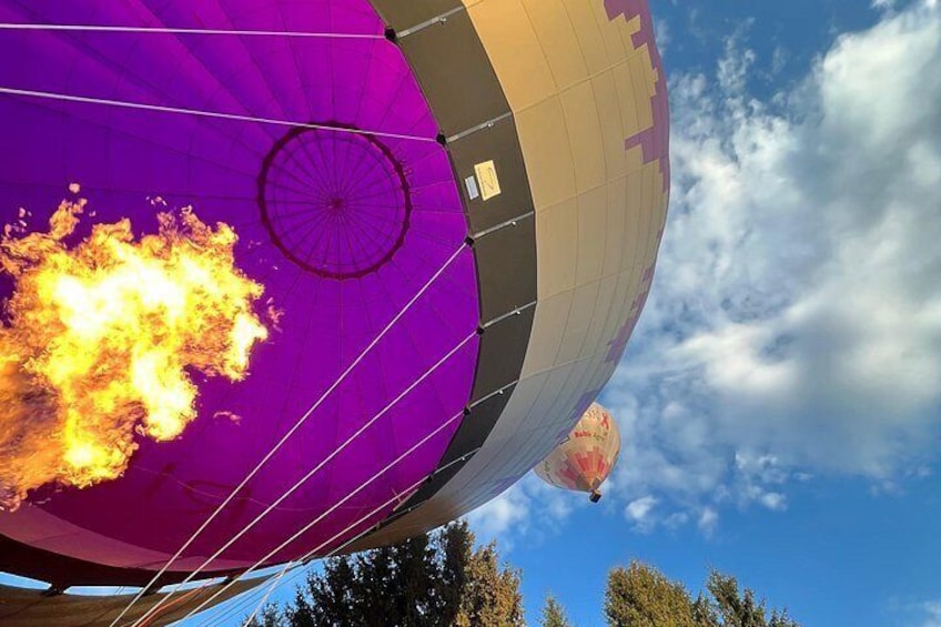 A hot air balloon with a purple and beige color scheme, a visible burner flame, and a clear blue sky with clouds. 4
