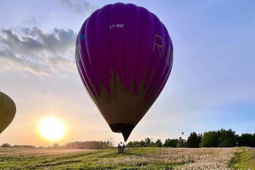 A purple hot air balloon with 'LY-RNT' and 'R' on it, flying over a field with the sun setting in the background. 2