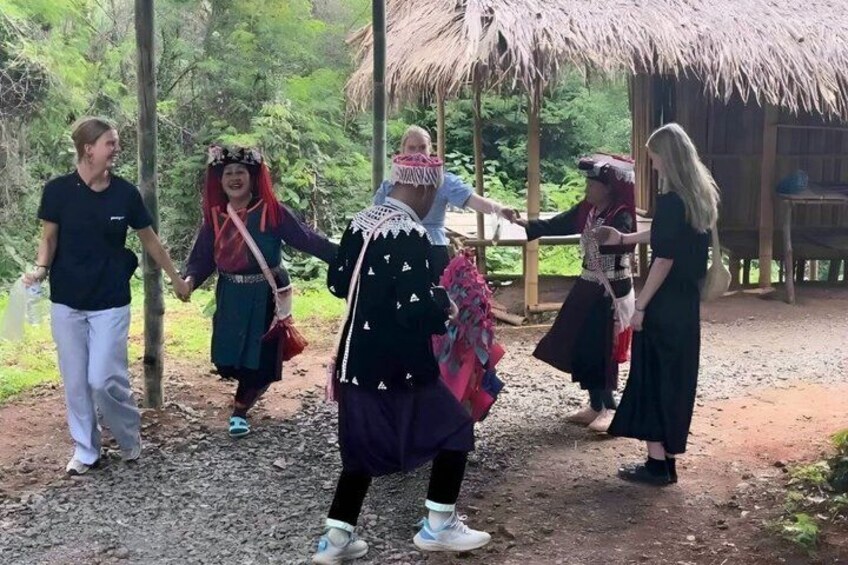 Un grupo de personas vestidas con atuendo tradicional caminan por un sendero de tierra en un área boscosa, con una estructura de techo de paja al fondo. 3