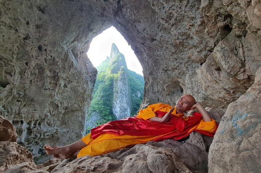 A person in orange robes sitting in a cave with a mountain in the background. 5