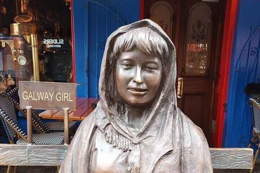 Une statue d’une femme, avec une pancarte sur laquelle on peut lire « GALWAY GIRL », se trouve devant un bâtiment dont les portes sont bleues et rouges. 4