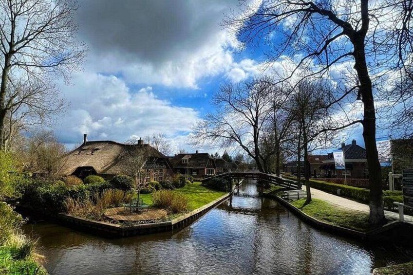 Un canal surmonté d’un pont, un bâtiment à toit de chaume et des arbres sans feuilles sous un ciel partiellement nuageux. 4