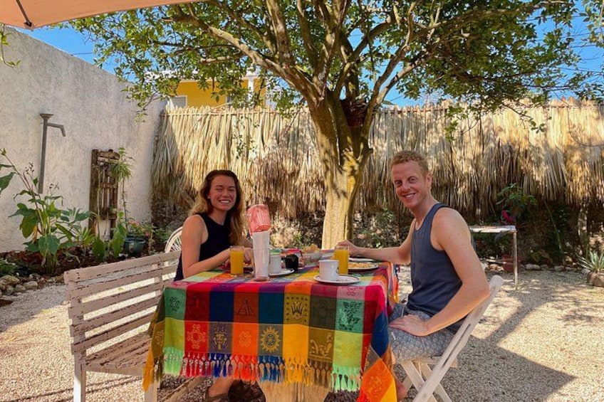 Two people sitting at a table with a colorful tablecloth, a tree, and a thatched roof in the background. 2