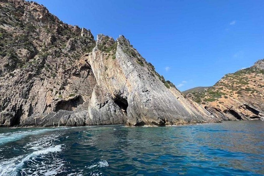 A rocky coastline with a clear blue sea, a prominent cliff, and a small cave-like opening. 2