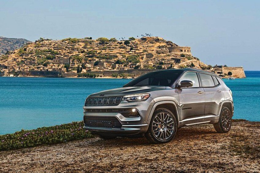 En silverfärgad SUV står parkerad på en stenstrand med en kustlandskap och historiska byggnader i bakgrunden. 2