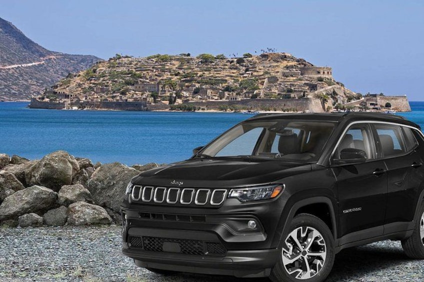 En svart SUV står parkerad på en stenig strand, med en kuststad och berg i bakgrunden. 3