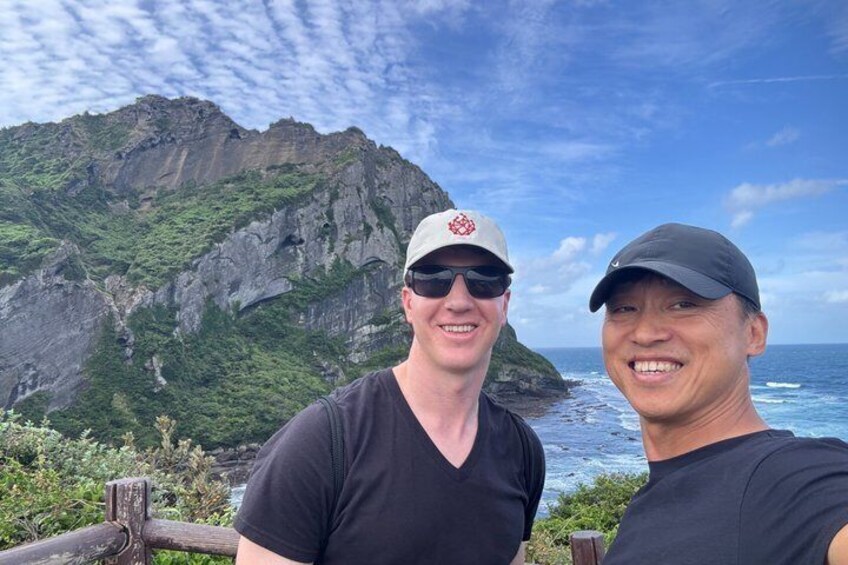 Two people standing in front of a mountainous landscape with a view of the ocean. 1