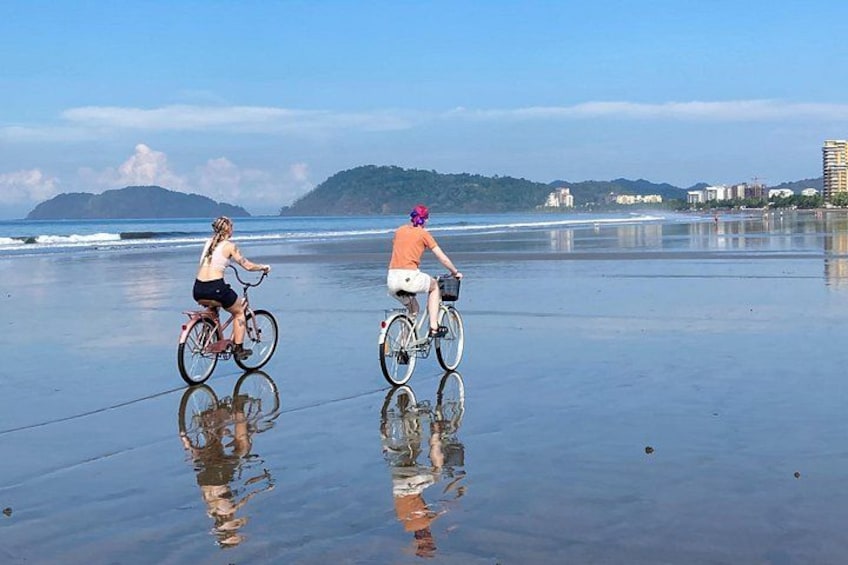 Two people riding bicycles on a wet beach with ocean and hills in the background. 3
