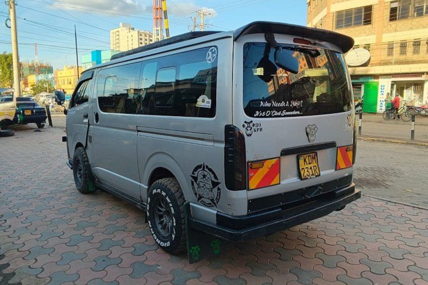 A silver van parked on a paved area with a license plate, surrounded by buildings and other vehicles. 4