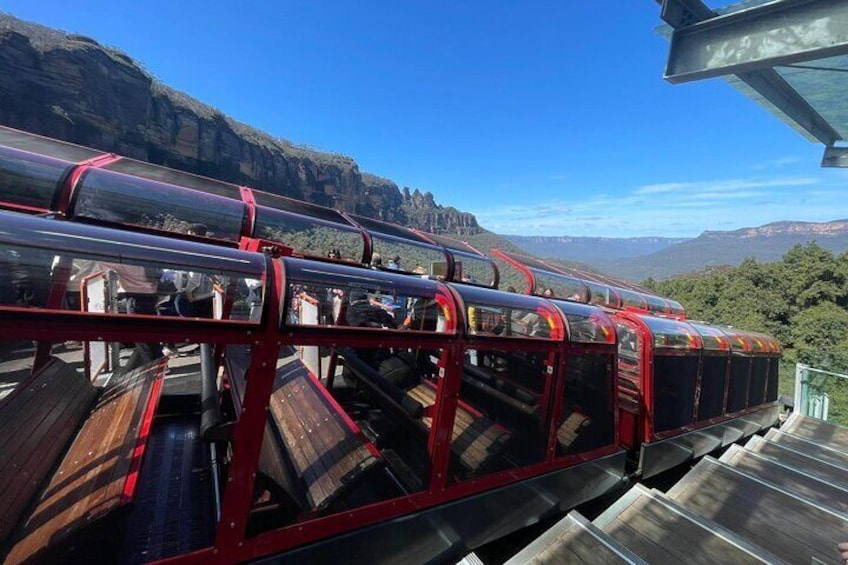 A red and black roller coaster with wooden seats, set against a mountainous landscape. 1