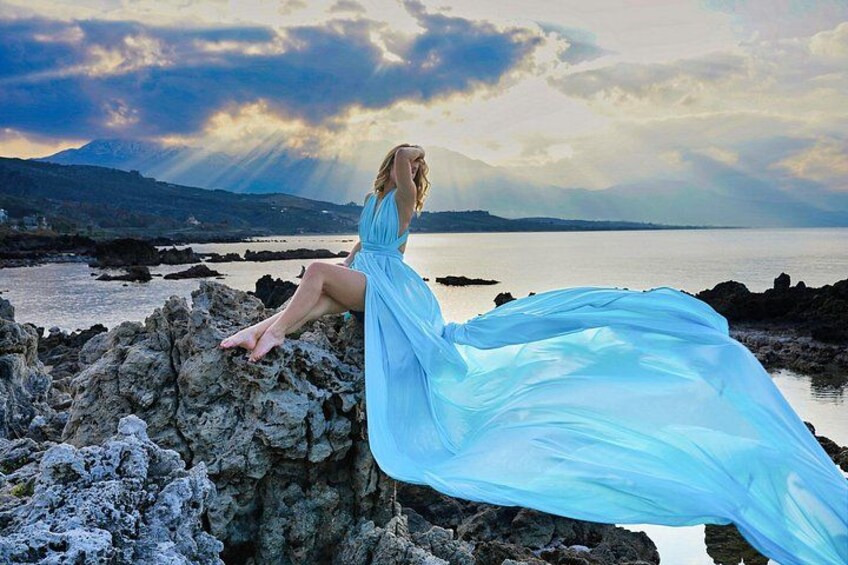 A person in a flowing blue dress sitting on rocks by a body of water with mountains in the background. 3