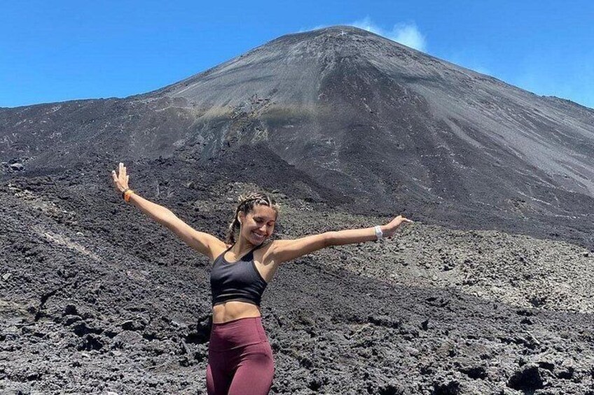 Una persona de pie sobre terreno rocoso, con los brazos levantados, frente a un volcán imponente. 4