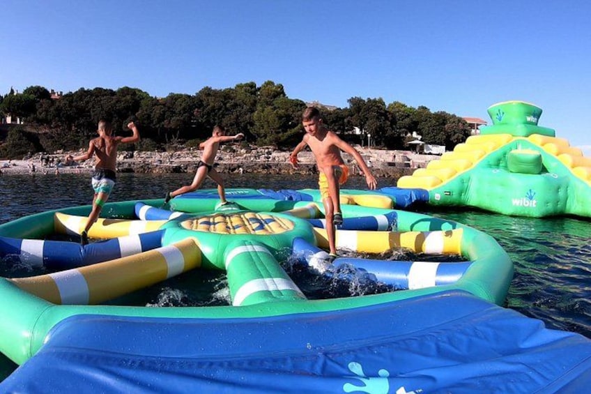 People playing on inflatable water structures in a body of water with a beach and trees in the background. 3