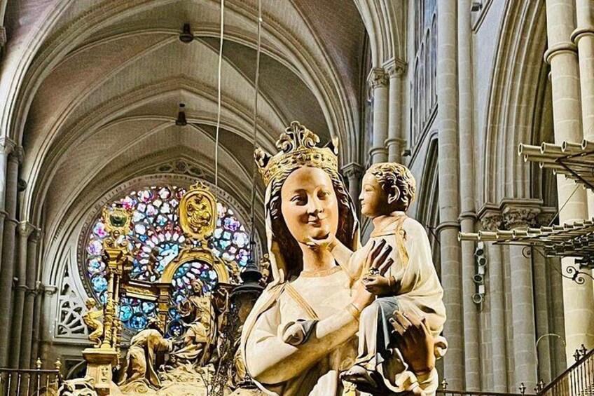 En statue af en kronet figur der holder et barn i en katedral med et farvet glasvindue i baggrunden. 5