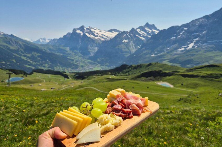 A wooden board with cheese, grapes, and cured meats against a mountainous backdrop. 1